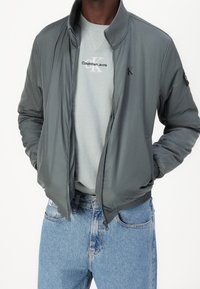 Bomberjacke aus hellgrauem Stoff mit gerippten Bündchen und Kragen; über einem hellblauen Pullover getragen und kombiniert mit hellblauen Jeans.