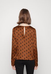 Femme aux cheveux bruns courts portant un chemisier marron à manches longues avec des pois noirs et une ouverture en forme de trou de serrure dans le dos, assorti à un pantalon noir.