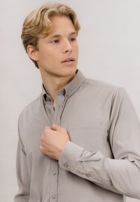 Lysegrå skjorte med knapper laget av myk stoff. Har standard krage, en enkelt brystlomme og knappede ermer. Glatt tekstur.