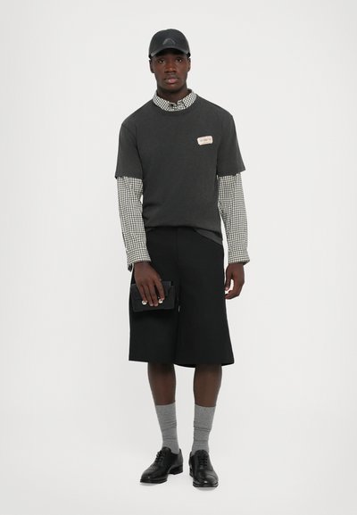 Homem a usar um boné preto, camisa de manga curta cinza escura por cima de uma camisa de manga longa xadrez, calções pretos até ao joelho, meias cinzas, sapatos de dress preto, segurando uma bolsa preta.