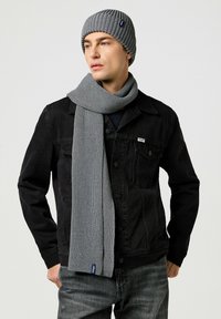 Beanie grigia in maglia e sciarpa abbinata con texture a coste, abbinati a una giacca di jeans nera e jeans grigi. Presenta un piccolo logo sulla sciarpa.