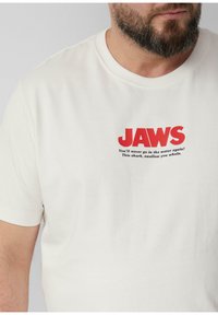 Lichtcreme korte mouwen t-shirt van zachte katoen. Heeft vet rood tekst "JAWS" en een slogan in kleinere zwarte letters eronder.