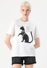 Hvit bomulls t-skjorte med grafikk av en svart katt med detaljer av perlekjede og tekst. Designet er plassert sentralt på brystområdet.