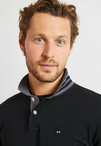 Homme avec des cheveux courts et bouclés et une barbe, portant une chemise noire à col avec un col intérieur rayé, regardant droit devant lui sur fond uni.