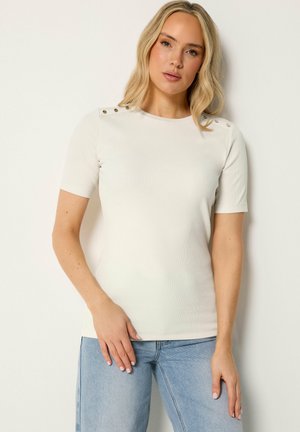 Blonde Frau trägt ein weißes kurzärmeliges geripptes Oberteil mit goldenen Knöpfen auf den Schultern und hellblaue Jeans, steht vor einer schlichten Wand.