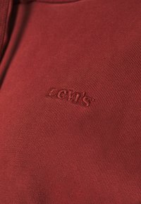 Tessuto rosso scuro con una texture a coste, caratterizzato da un logo "Levi's" in rilievo. Il design è semplice e aderente.