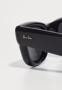Black Ray-Ban sunglasses feature a glossy plastic frame, grey lenses, and a gold logo accent on the right temple.