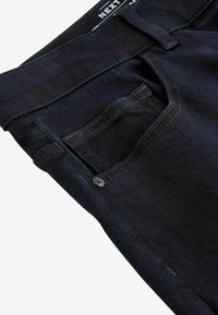 Jeans de mezclilla negra con un bolsillo delantero con botón, costuras visibles y una textura suave. Etiqueta de la marca ubicada en la cinturilla.