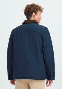 Padded jakna v temno modri barvi s žametnim ovratnikom, ki ima gumbaste manšete, ravno spodnji rob in teksturo v obliki diamanta na tkanini.