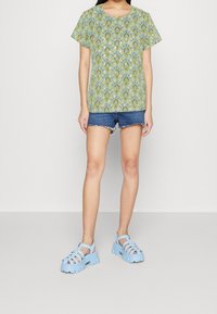 Haut à motifs verts à manches courtes avec un design floral, associé à un short en denim bleu et des sandales chunky bleu clair.