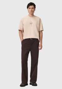 Camiseta de algodón beige con mangas cortas y diseño de texto centrado. Combinada con pantalones marrones oscuros y zapatos negros. Look sencillo y casual.