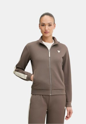 Donna che indossa una giacca sportiva marrone con zip e pantaloni abbinati con strisce bianche ai lati, in piedi con una mano in tasca su uno sfondo neutro.