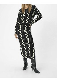 Robe tricotée à motifs noir et blanc avec des manches longues, présentant des designs ondulés et une silhouette ajustée, associée à des bottes noires jusqu'aux genoux.