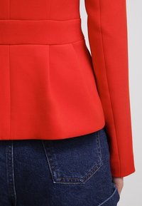 Blazer rouge ajusté avec un ourlet structuré porté sur un jean en denim bleu foncé, montrant le dos et le côté droit de la tenue.