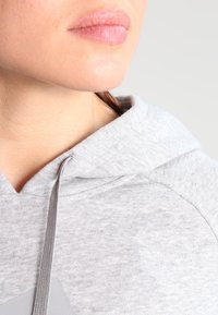 Sudadera gris con capucha de tela texturizada, que cuenta con capucha con cordón y un detalle de costura visible cerca del cuello.