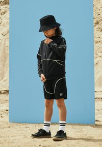 Enfant portant un chapeau bob noir, une veste noire et un short avec un liseré blanc, debout sur un sol sablonneux devant un fond bleu.