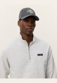 Grå zip-up hettegenser med svart logo-patch og matchende grå cap med grønn brodert tekst, båret av en person.