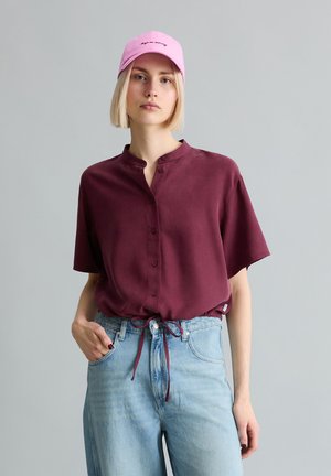 Jeune femme portant une casquette rose, un chemisier bordeaux à manches courtes avec boutons, et un jean taille haute bleu clair, une main dans la poche.
