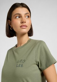 Oliwkowa bawełniana koszulka z krótkim rękawem i okrągłym dekoltem; na piersi nadrukowane symbole pokoju i ręki oraz napis "LEE".