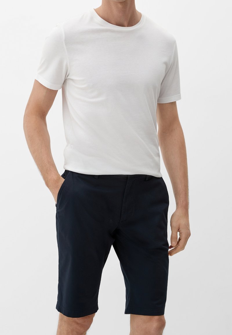 Homme portant un t-shirt blanc uni à manches courtes et un short bleu marine foncé arrivant aux genoux, mains détendues le long du corps.