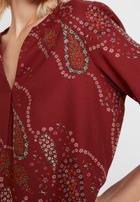 Blouse rouge avec un col en V, manches courtes, ornée de motifs floraux paisley en blanc, vert et rose sur un tissu texturé.