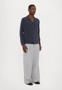 Marineblaues Button-Up-Hemd mit einer Tasche, kombiniert mit lockeren, hellgrauen Hosen und schwarzen Schuhen. Glattes Gewebe mit leichtem Glanz.