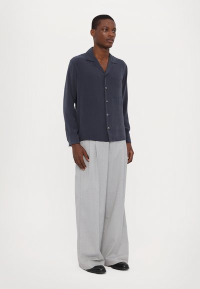 Camisa de botón azul marino con un bolsillo, combinada con pantalones sueltos de un gris claro y zapatos negros. Tejido suave con un ligero brillo.