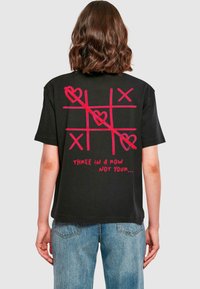 Zwarte katoenen t-shirt met een grote rode tic-tac-toe graphic met harten en de tekst "DRIE OP EEN RIJ NIET JOUW..." op de achterkant.