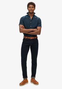 Camicetta corta blu scuro, cintura marrone, jeans scuri e stivali marroni con lacci. La camicia ha un colletto con bottoni; i jeans sono slim-fit.