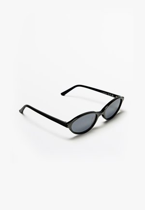Lunettes de soleil ovales noires avec monture fine et verres teintés foncés, posées sur une surface blanche avec une ombre en dessous.