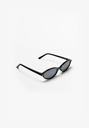 Lunettes de soleil ovales noires avec monture fine et verres teintés foncés, posées sur une surface blanche avec une ombre en dessous.