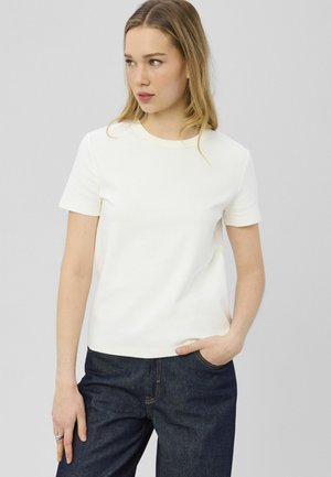 Femme aux longs cheveux blonds portant un t-shirt blanc uni à manches courtes et un jean bleu foncé, debout avec une main dans la poche.