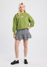 Groene fleece sweatshirt met rond logo, gecombineerd met een zwart-witte gingham roezelrok. Zwarte instappers en crèmekleurige sokken maken de look compleet.
