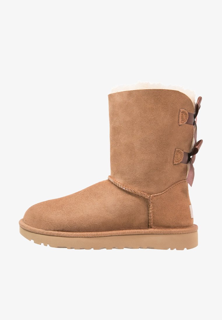 Beige Wildleder-Ankle-Boot mit weicher weißer Fellfütterung, dekorativen braunen Riemen und Schleifen an der Rückseite sowie einer flachen Gummisohle für Komfort.