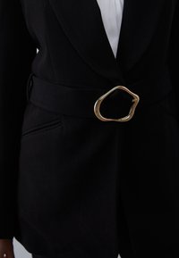 Zwarte op maat gemaakte blazer met brede bijpassende riem met onregelmatige gouden gesp, gedragen over een wit topje.