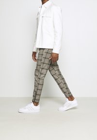 Giacca bianca con due tasche sul petto, pantaloni a quadretti beige e marroni, e sneakers bianche con tre righe di accentuazione, indossati da una persona in piedi.