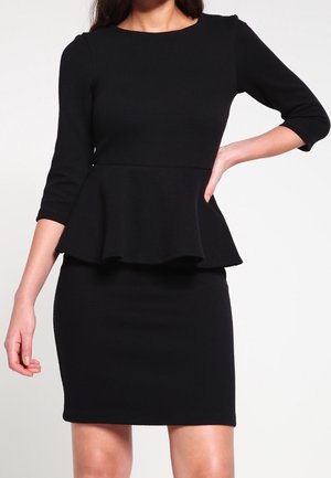 Kobieta ubrana w czarną sukienkę peplum z rękawami do łokcia, dopasowaną w talii i spódnicą sięgającą kolan na jednolitym tle.