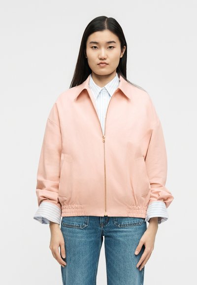 Hosszú sötét hajú nő világos rózsaszín cipzáras dzsekit visel fehér csíkos ing fölött és kék farmert, egyszerű háttér előtt áll.