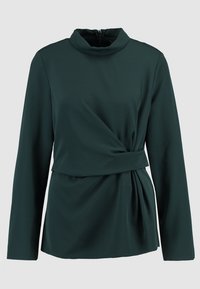 Tmavě zelený top s dlouhým rukávem a vysokým límcem, s překříženým předním designem a strukturovanou látkou, která poskytuje hladký, splývavý vzhled.