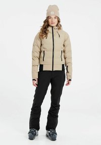 Beige gepolsterte Jacke mit Reißverschluss vorne, schwarzen Akzenten und Kapuze, kombiniert mit schwarzen Skihosen und Stiefeln. Beige gestrickte Mütze auf dem Kopf.