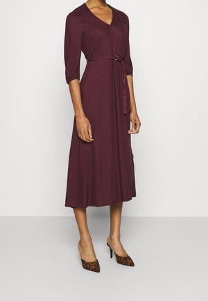 Vrouw draagt een knielange pruimkleurige wikkeljurk met een tailleceintuur en driekwart mouwen, gecombineerd met hoge hakken met luipaardprint.