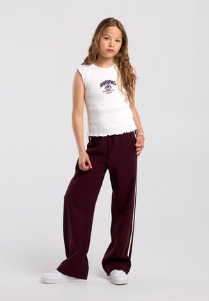 Meisje met lang haar, gekleed in een witte mouwloze top, bordeauxrode wijde broek met zijstrepen en witte sneakers, staand tegen een effen achtergrond.