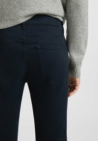 Pantaloni in denim blu scuro con due tasche posteriori, cuciture a contrasto e una texture liscia, indossati con un maglione lavorato a maglia grigio chiaro.