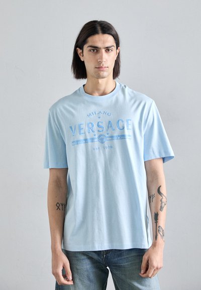 Himmelblaues Baumwoll-T-Shirt mit einem grafischen Druck, der "MILANO VERSACE" in einem kreisförmigen Design zeigt. Kurze Ärmel und eine lockere Passform.