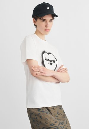 Mladý člověk s černou čepicí a bílým tričkem Carhartt WIP, který má zkřížené ruce, stojící před jednobarevným světlým pozadím.