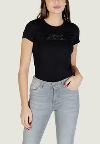 Femme portant un t-shirt noir ajusté à manches courtes avec un logo "Armani Exchange" en strass et un jean taille haute gris clair, main dans la poche.
