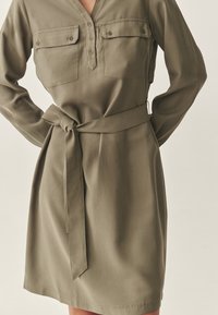 Olivgrünes Kleid mit Gürtel, V-Ausschnitt, langen Ärmeln und zwei Brusttaschen. Aus weichem Stoff mit glatter Textur und taillierter Passform.