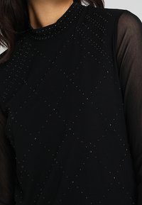 Top nero con collo alto, tessuto testurizzato e decorazioni a diamante con borchie. Maniche lunghe e trasparenti si estendono dalle spalle.