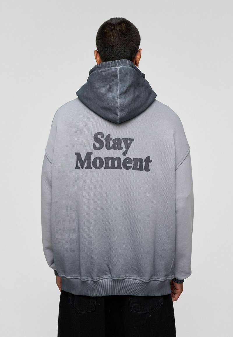 Grijze hoodie met een zwarte "Stay Moment" print op de achterkant. Het heeft een capuchon met trekkoord en ribgebreide boorden en zoom. Glad textuur van de stof.