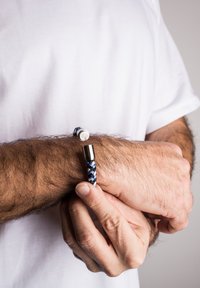 Zils un balts pītais rokassprādze ar sudraba aizdari, kurā ir apaļš logotipa elements. To nēsā uz rokas ar redzamiem matiem un roku.